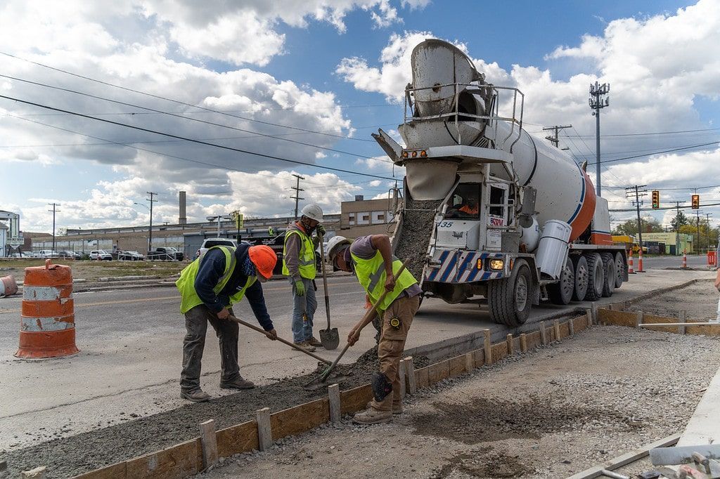 Construction workers pouring concrete from a mixer truck on a road under cloudy skies