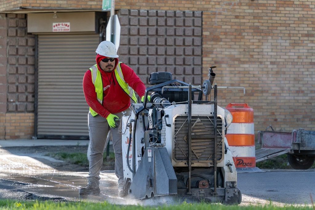 Road worker in red vest operating pavement equipment near a building and orange construction barrel