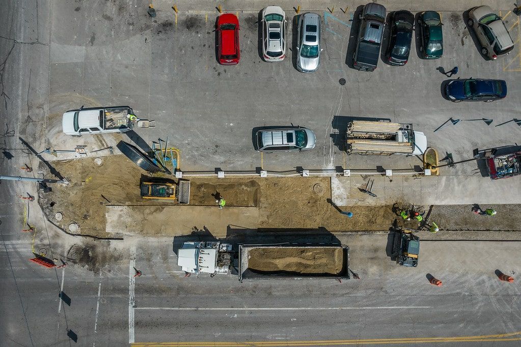Aerial view of a roadwork site with excavators, a trench, and parked cars along the street.