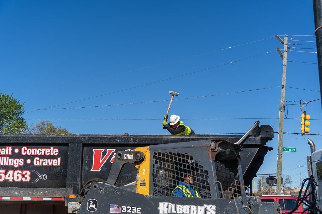 Worker on a black dump truck at a construction site under a clear blue sky.