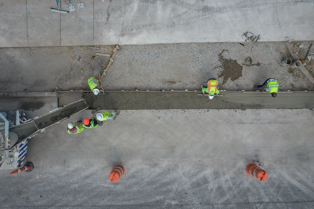 Construction workers in neon vests inspect a trench beside a concrete wall and sidewalk.