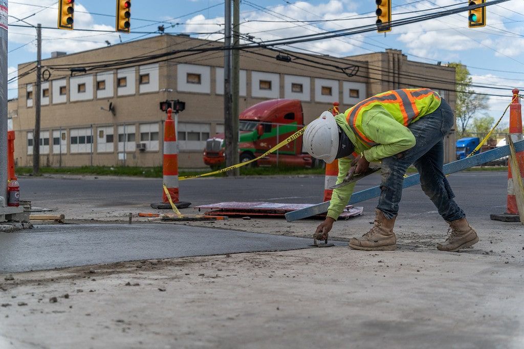 Road worker in a reflective vest shoveling debris at a wet intersection near traffic lights and a brick building