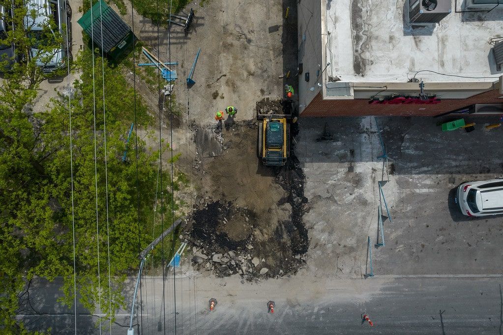 Overhead view of a street worksite with excavator, dirt pile, trees, and parked cars beside a building