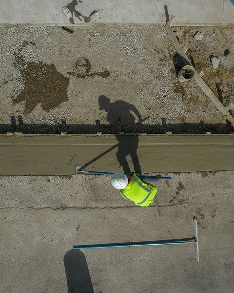 Worker in neon vest below a concrete ledge, with long shadow and tools on a rooftop.