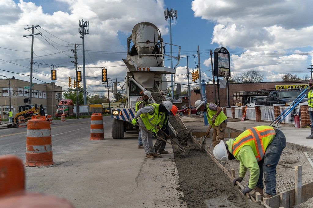 Construction workers paving a road at a busy intersection with orange traffic barrels and heavy equipment