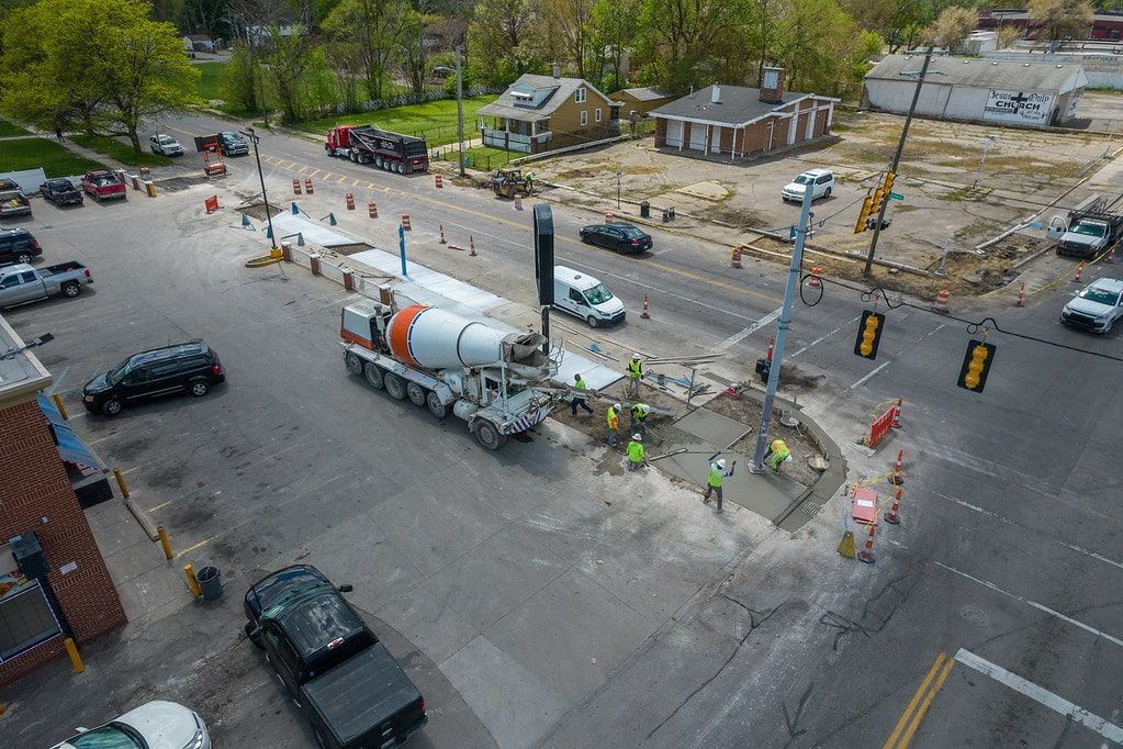 Street intersection with traffic cones, a cement truck, workers, and several cars near a wooded area.