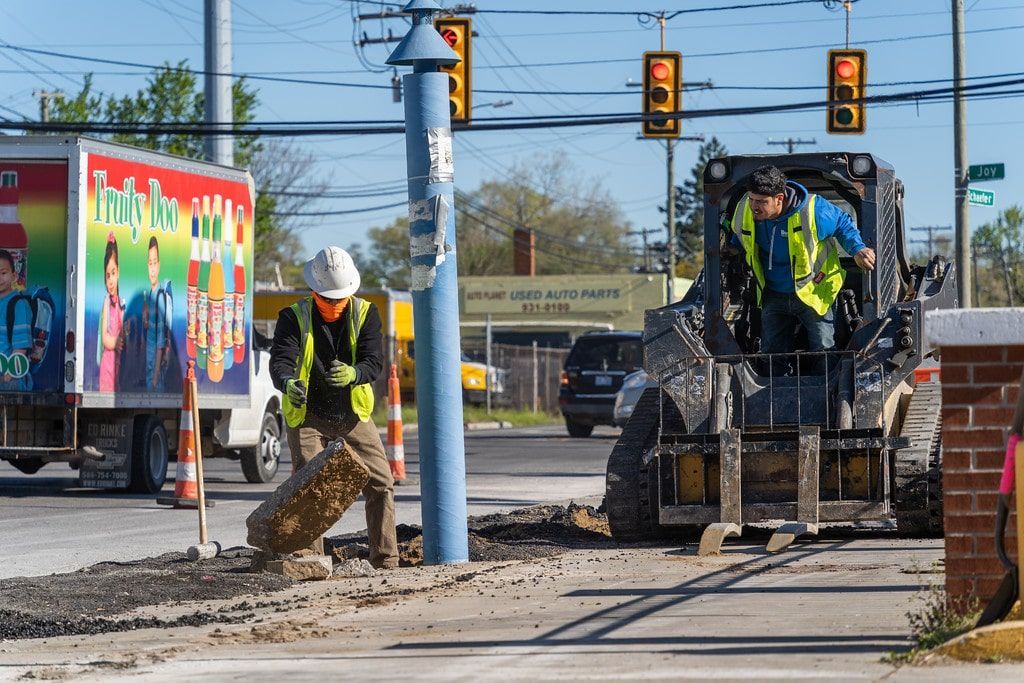 Road workers repairing a sidewalk beside a street, with a utility truck and traffic lights in the background.