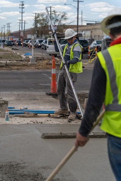 Road construction workers in reflective vests and hard hats pour concrete near a street.