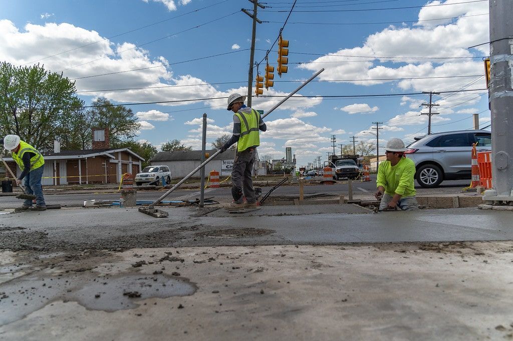 Road workers in neon vests pour concrete at a busy intersection under a cloudy sky.
