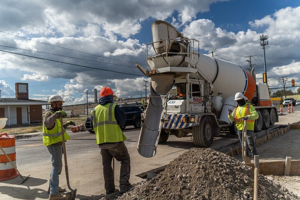 Construction workers in reflective vests guiding a cement mixer truck at a roadwork site under cloudy skies