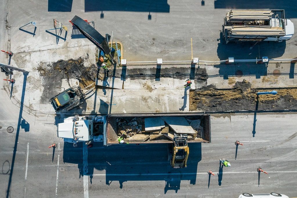 Overhead view of a road construction site with excavators, dump trucks, and workers on asphalt.