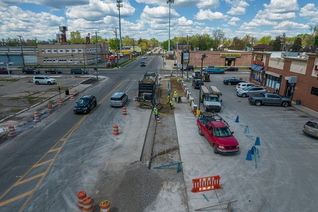 Roadwork on a wide city street with cars, orange barriers, and pedestrians under a cloudy sky
