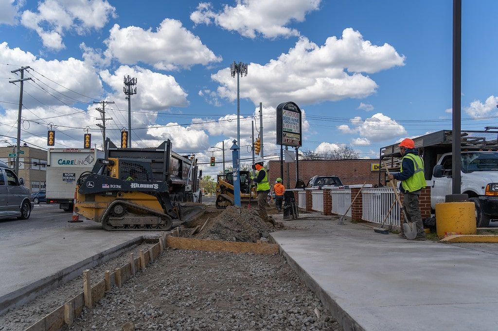 Roadwork with a skid steer spreading gravel beside a concrete path under a cloudy sky.