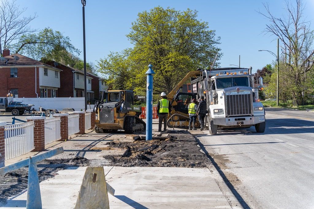 Road construction crew repairing a sidewalk beside a white truck on a sunny residential street