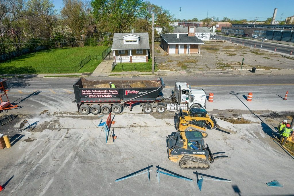 Construction site with excavators and workers on a paved area near buildings and a waterfront