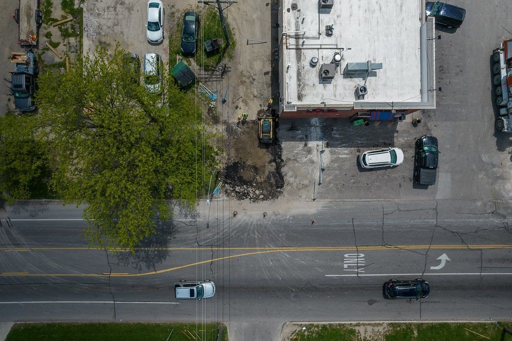 Aerial view of a street beside a building, with parked cars, trees, and a few vehicles on the road.