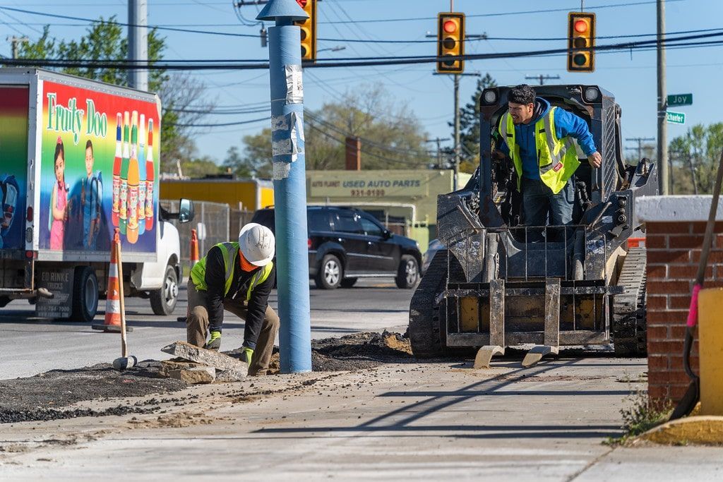 Road crew paving a sidewalk beside a colorful mural, with workers and a paving machine at an intersection.