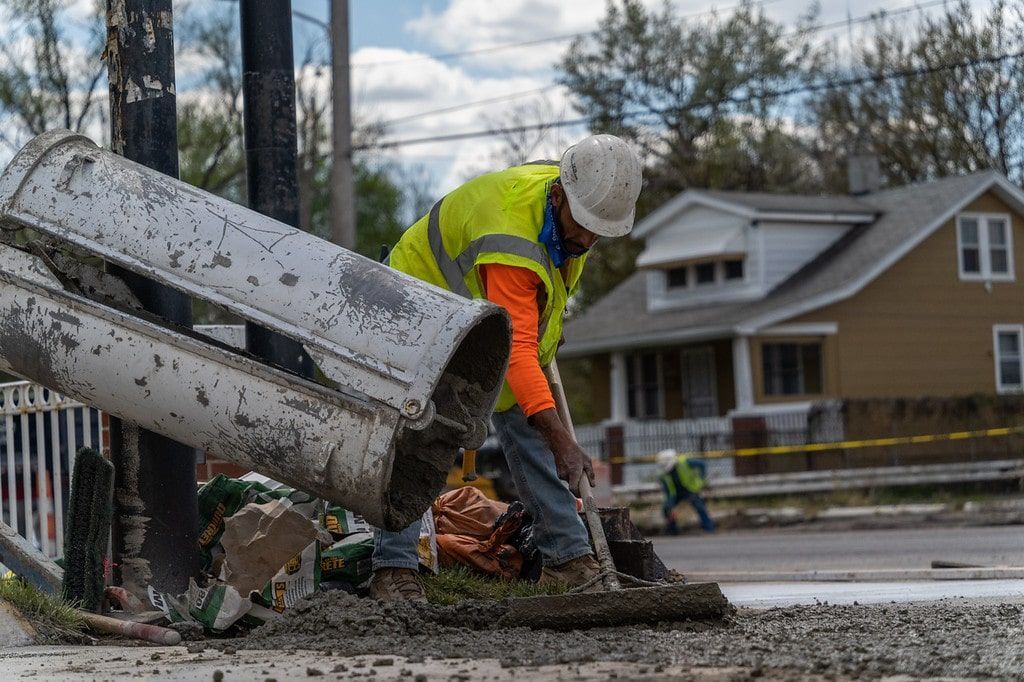 Worker in orange safety vest clears debris beside a damaged utility pole on a wet roadside.