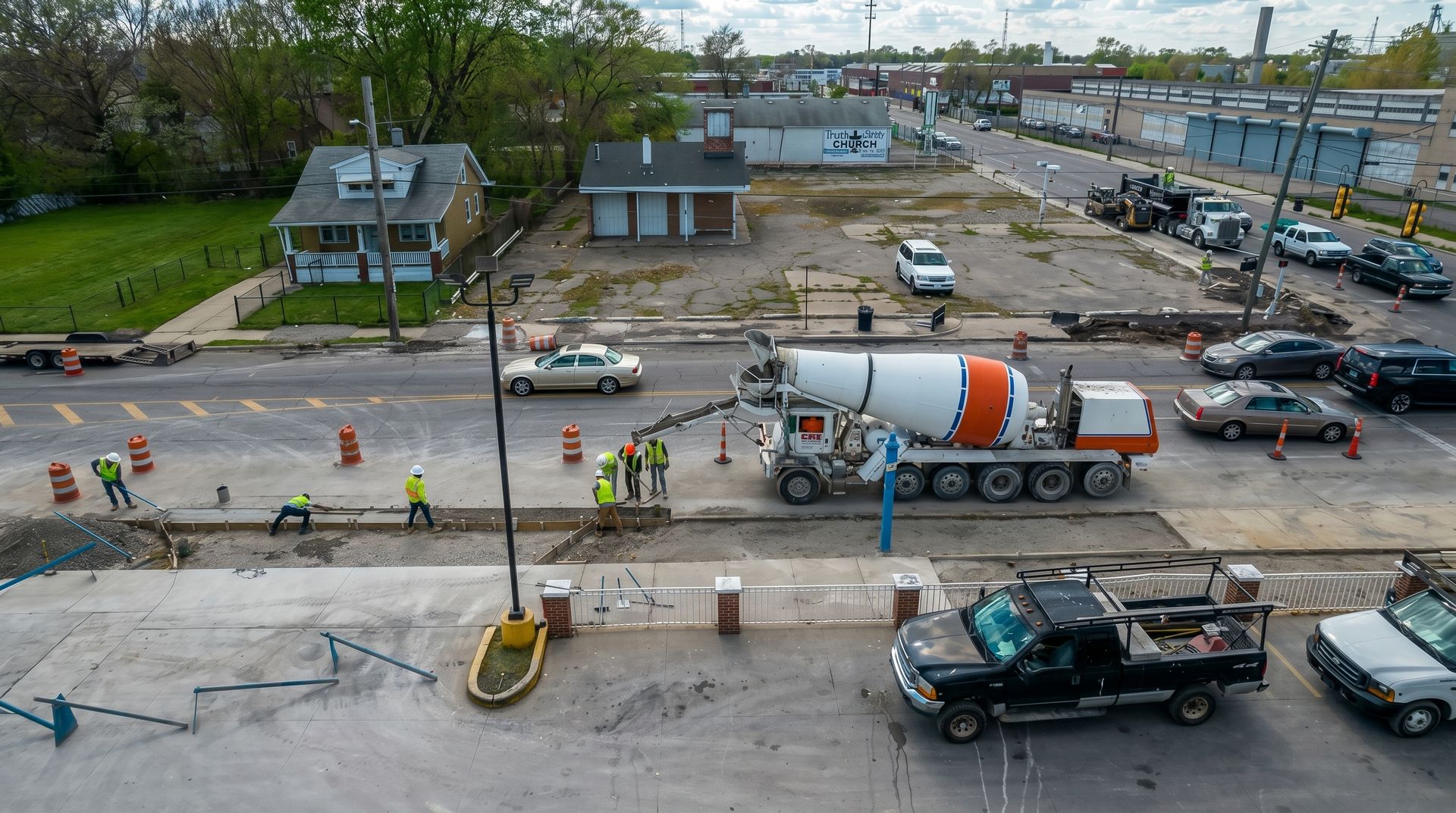 Construction site with workers, cement trucks, and road equipment on a paved lot near houses.