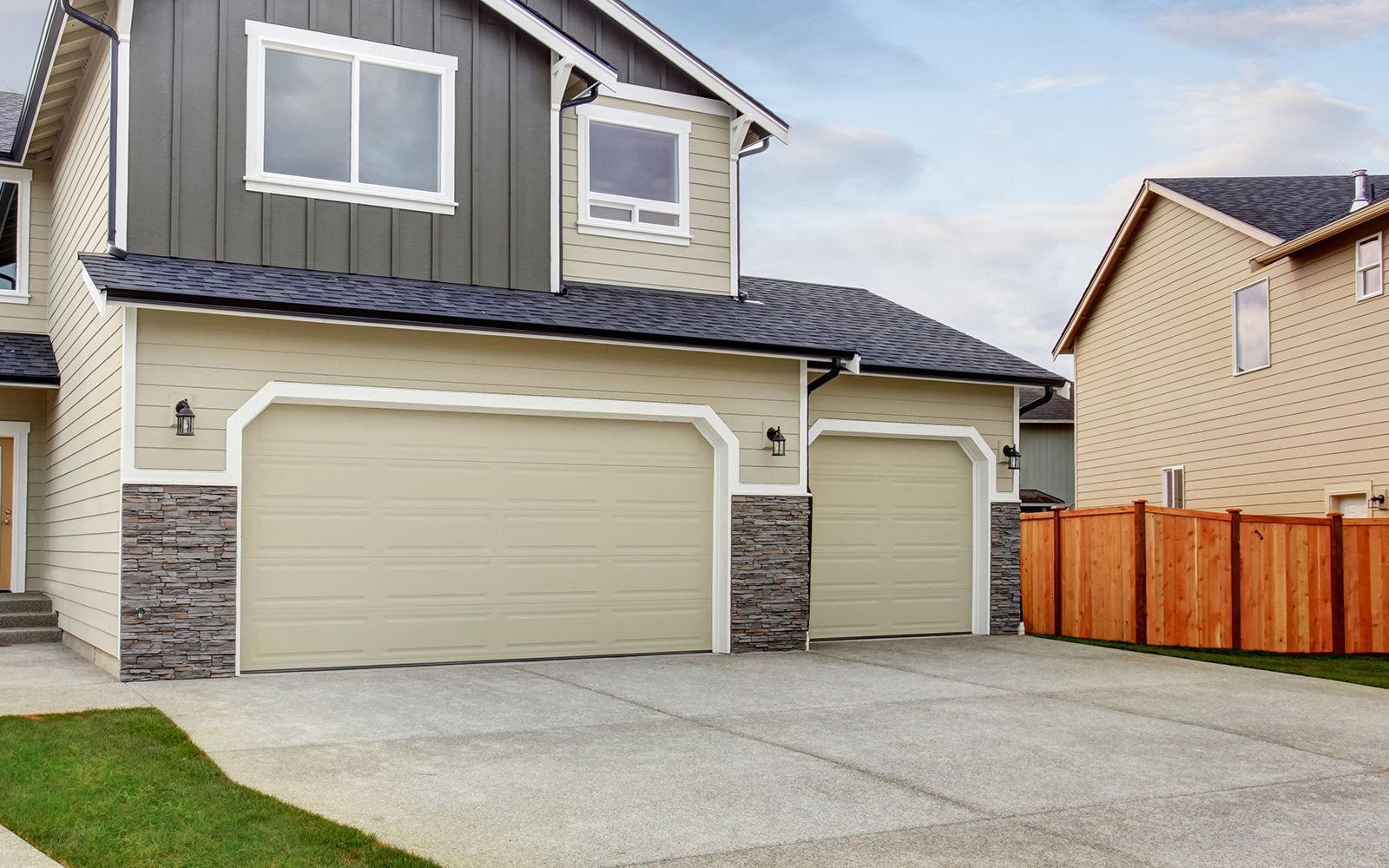 Suburban house with beige garage doors, stone accents, and a concrete driveway beside a wooden fence.
