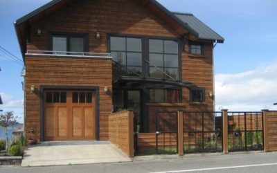 Two-story wooden house with large windows, front gate, and driveway under a blue sky