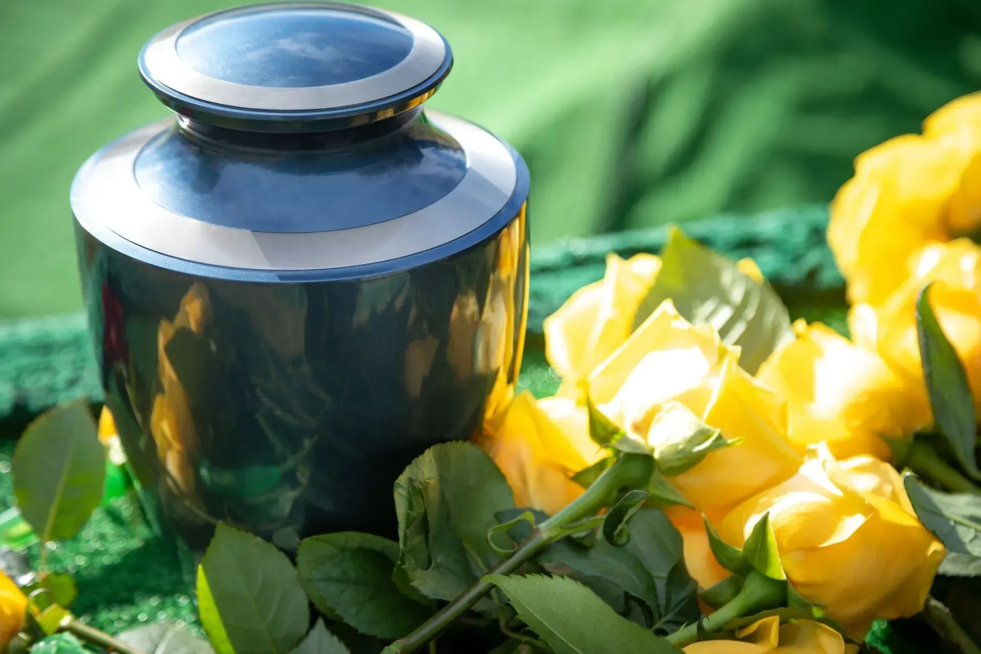 Blue urn with silver trim next to yellow roses on a green cloth.