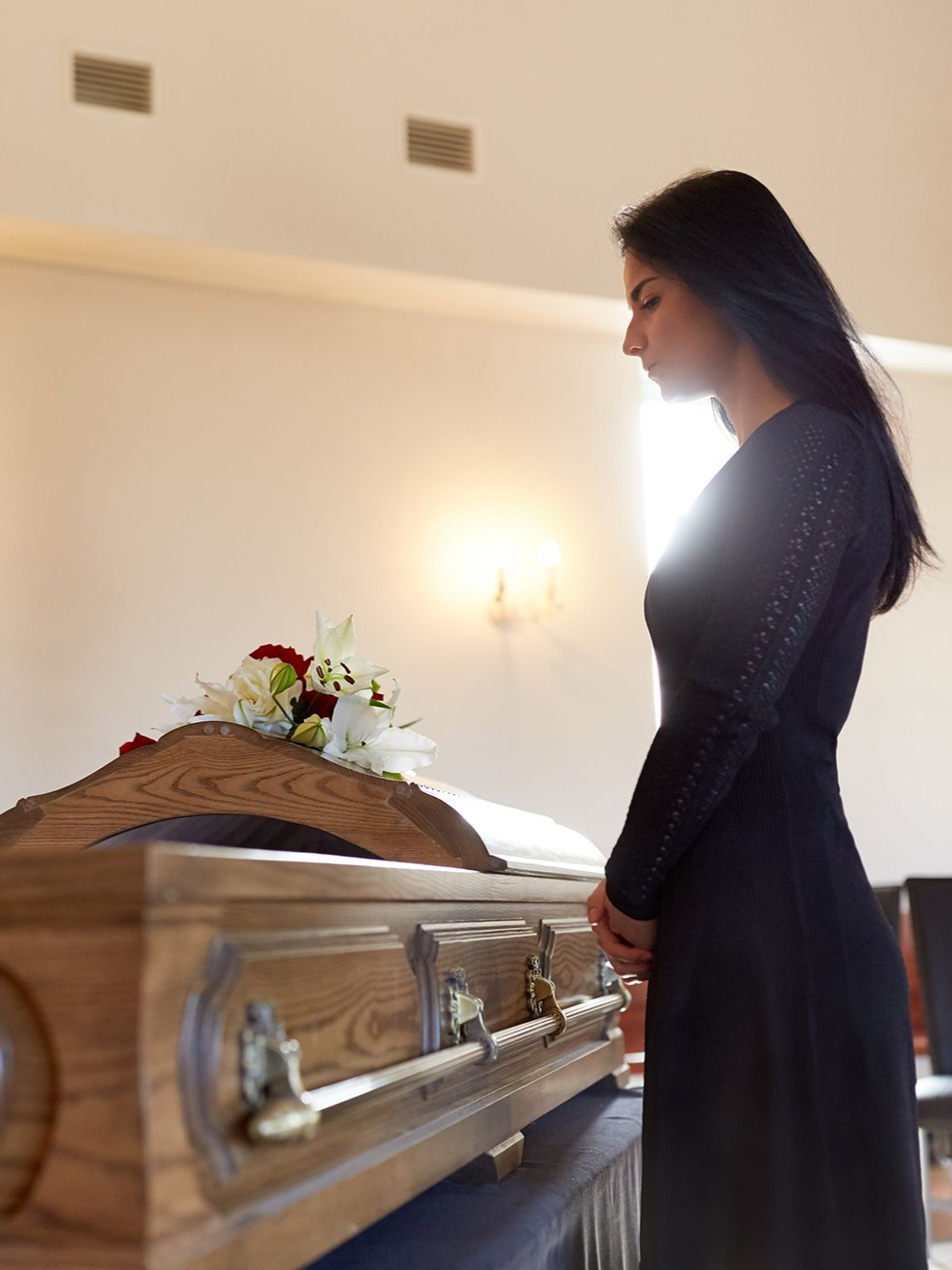 Woman in black dress at a funeral, looking down at a closed wooden casket with flower arrangement.