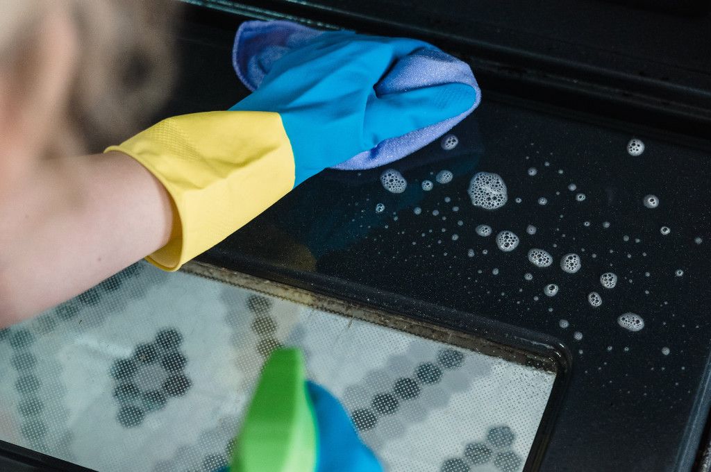 Cleaner cleaning an oven door with gloved hands and spray oven cleaner