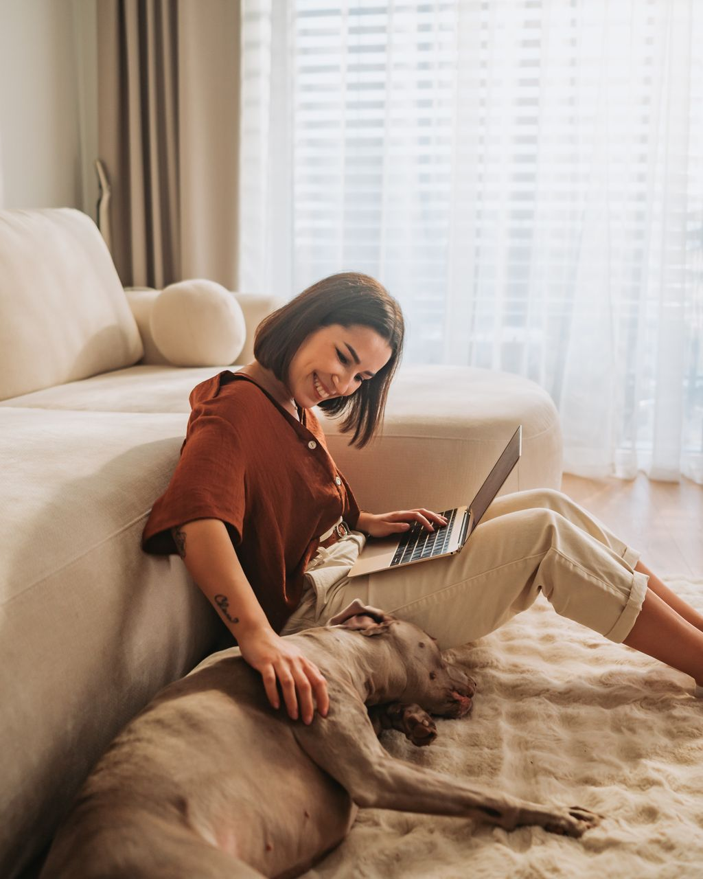 Woman smiles while using a laptop, petting a dog on a rug, sitting by a couch. Natural light floods the room.