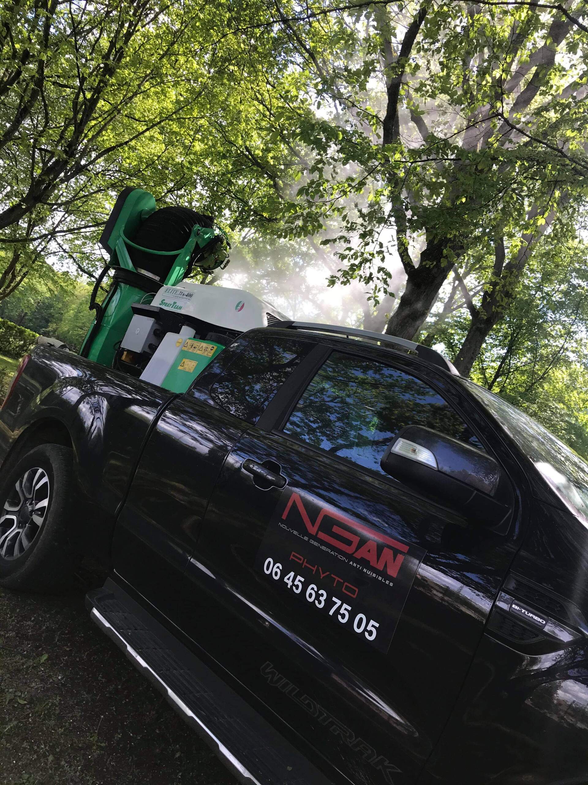 Un camion noir avec une machine verte à l'arrière est garé devant des arbres.