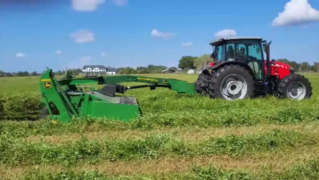 A red tractor pulling a green mower cutting tall grass in a field under a blue sky.