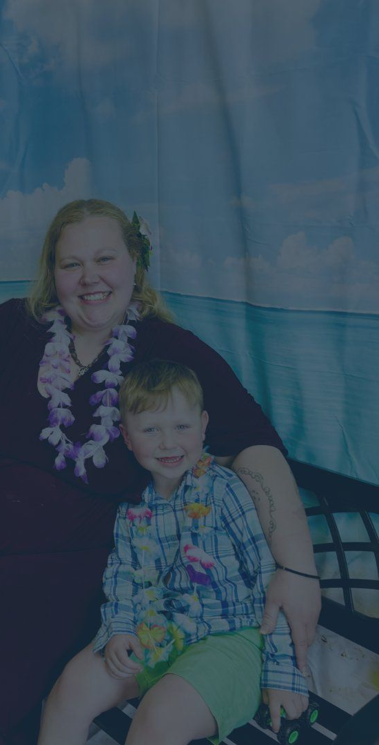 Woman and child sitting together, smiling, with a blue ocean backdrop and flower leis.