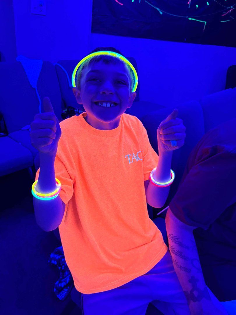 Child in a neon orange shirt smiling and giving two thumbs up under blue lighting, wearing glow bracelets.