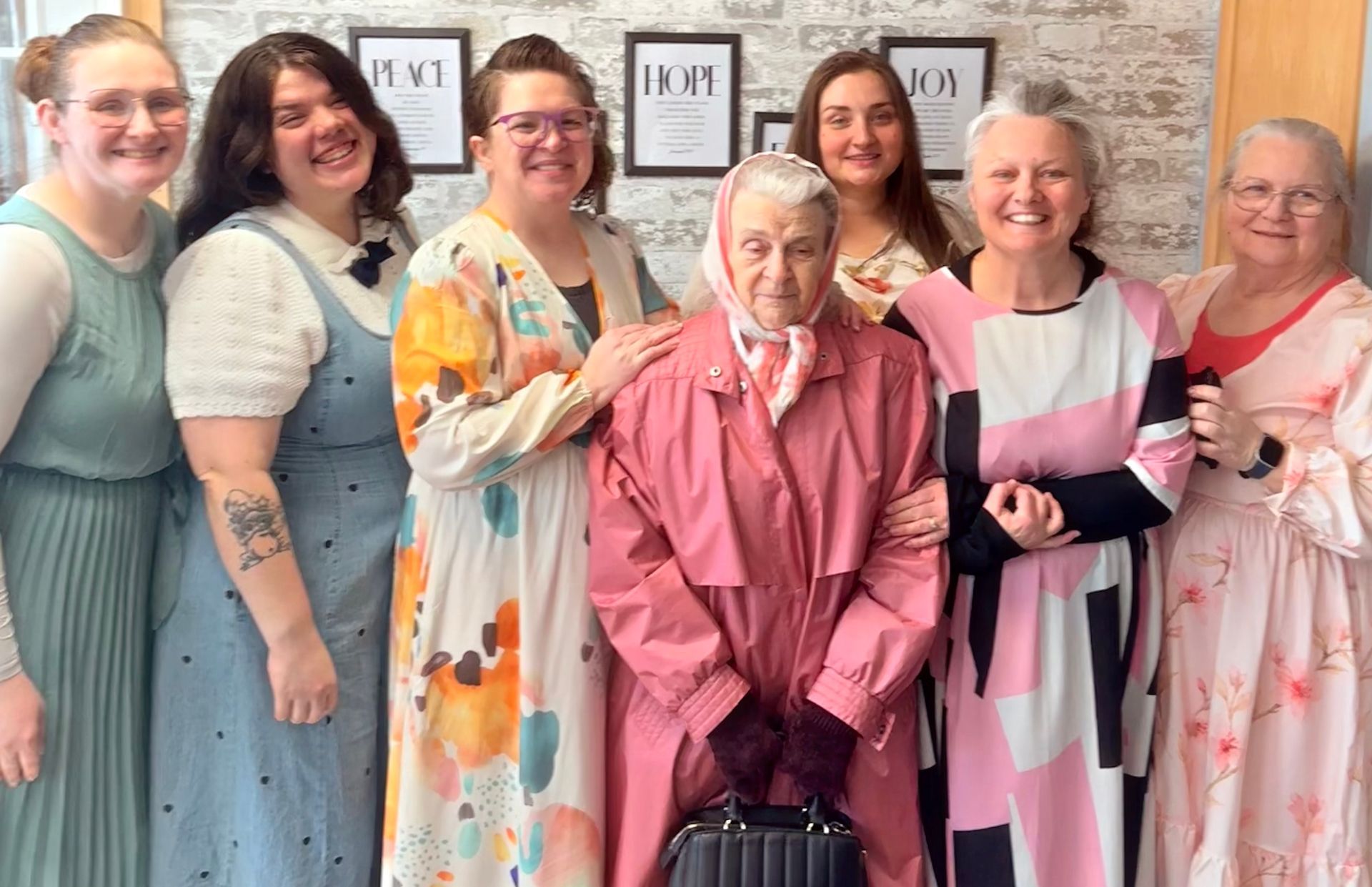Seven people pose together indoors in coordinated pink and floral outfits, smiling at the camera.