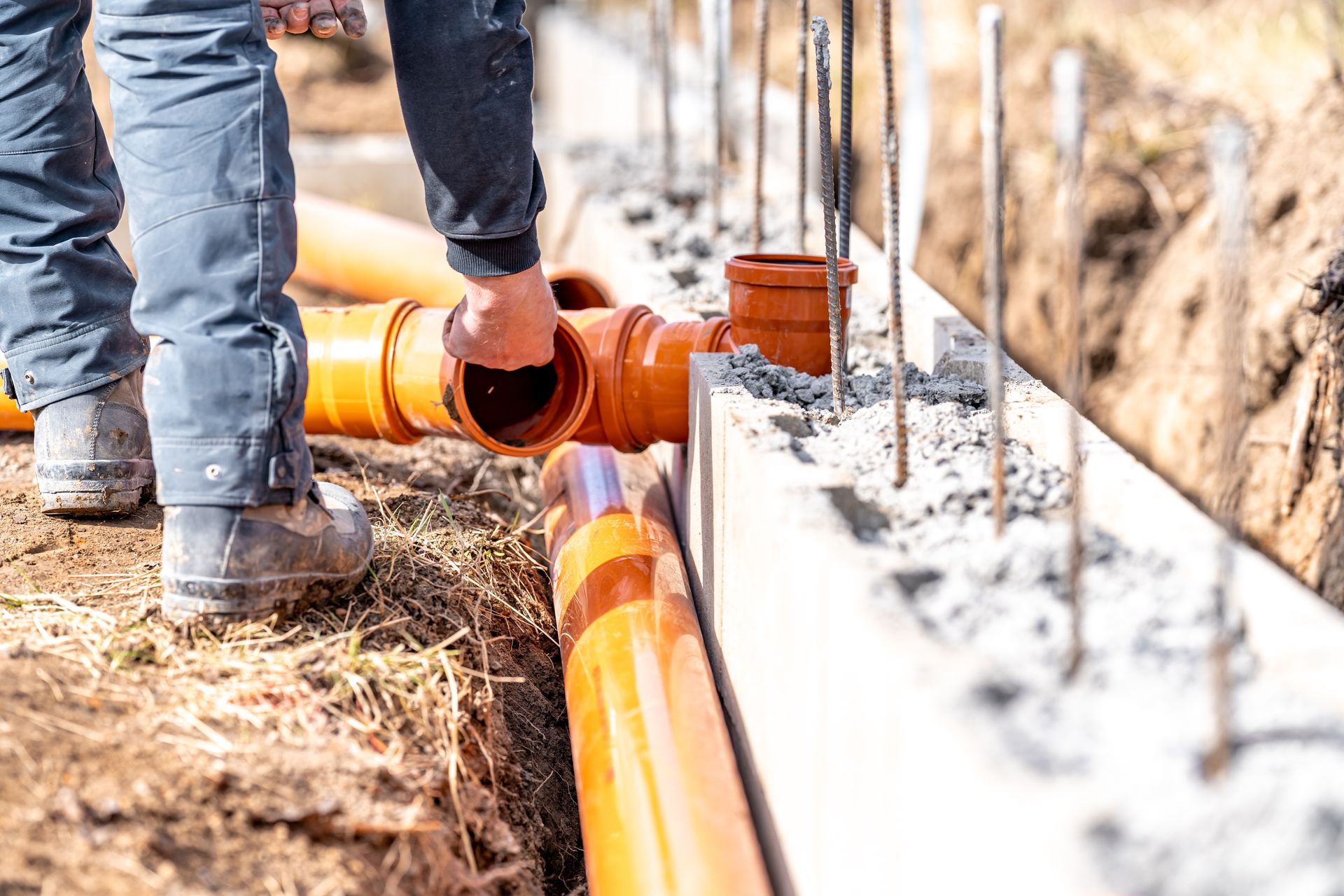 Construction worker connecting orange drainage pipes near a concrete foundation.