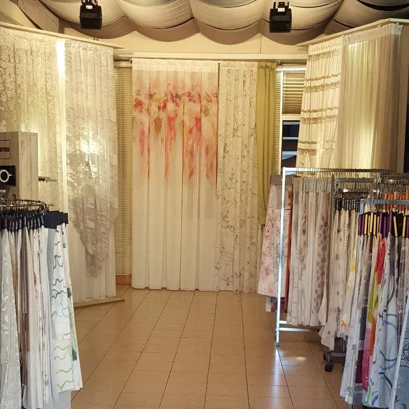 Corridoio di un negozio di abbigliamento con scaffali pieni di abiti e tende di colore chiaro, al centro di uno sfondo floreale bianco.