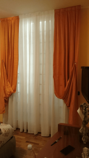 In un soggiorno, vicino a un televisore e ad alcuni mobili, tende bianche trasparenti incorniciate da drappeggi arancioni.