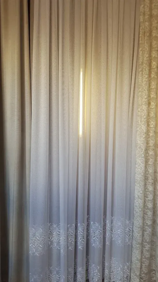 Tende beige trasparenti, con la luce del sole che filtra attraverso una stretta fessura al centro.