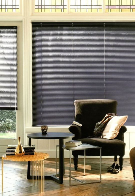 Soggiorno moderno con poltrona scura, tavolino, sgabello intrecciato e grandi finestre con tende.
