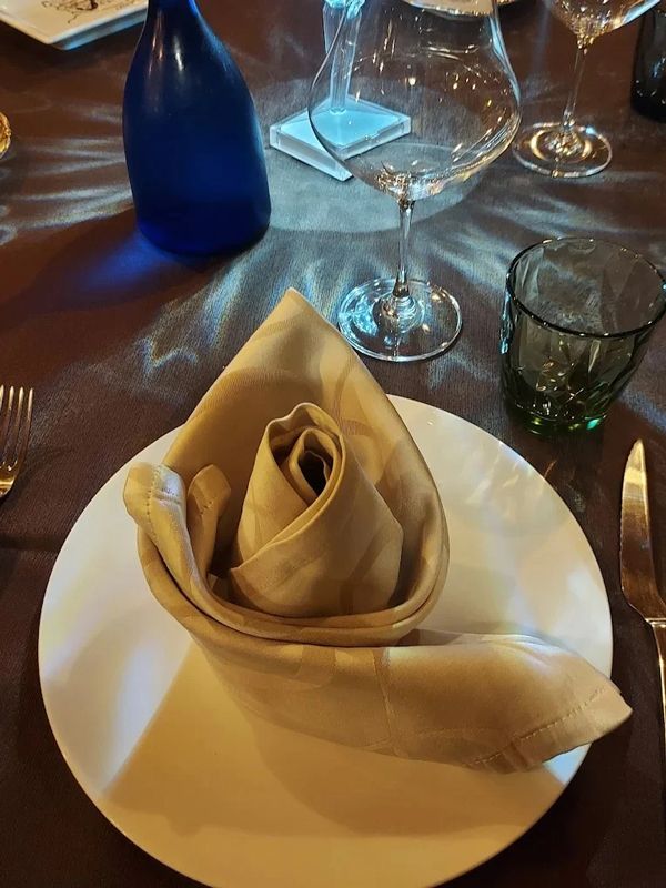 Un tovagliolo beige piegato a forma di rosa è appoggiato su un piatto bianco, su una tavola apparecchiata con bicchieri da vino e posate.
