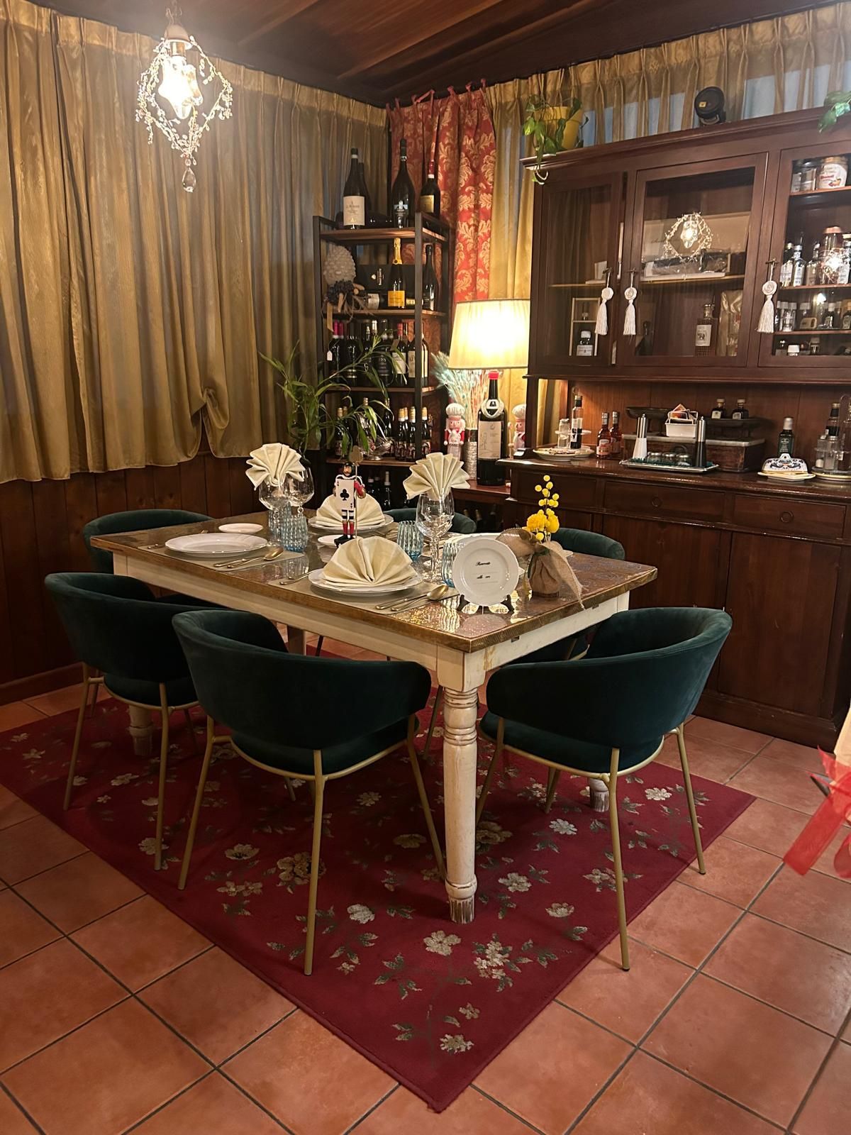 Una sala da pranzo con un tavolo dalle gambe bianche, sedie in velluto color verde acqua e un tappeto a motivi rossi su un pavimento piastrellato vicino a una credenza in legno.