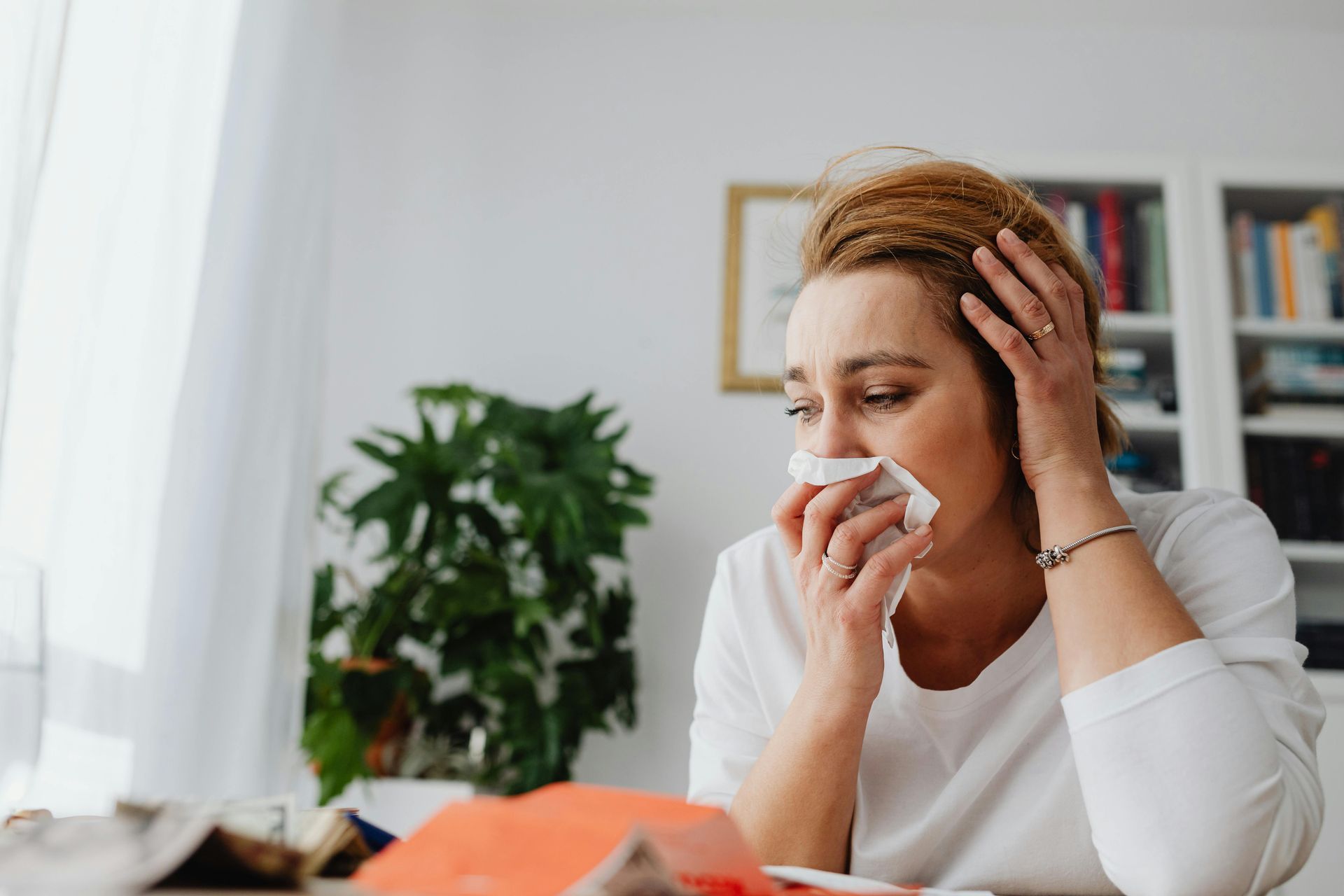 A person in a white sweater holding a tissue and their head, feeling tired from allergies.