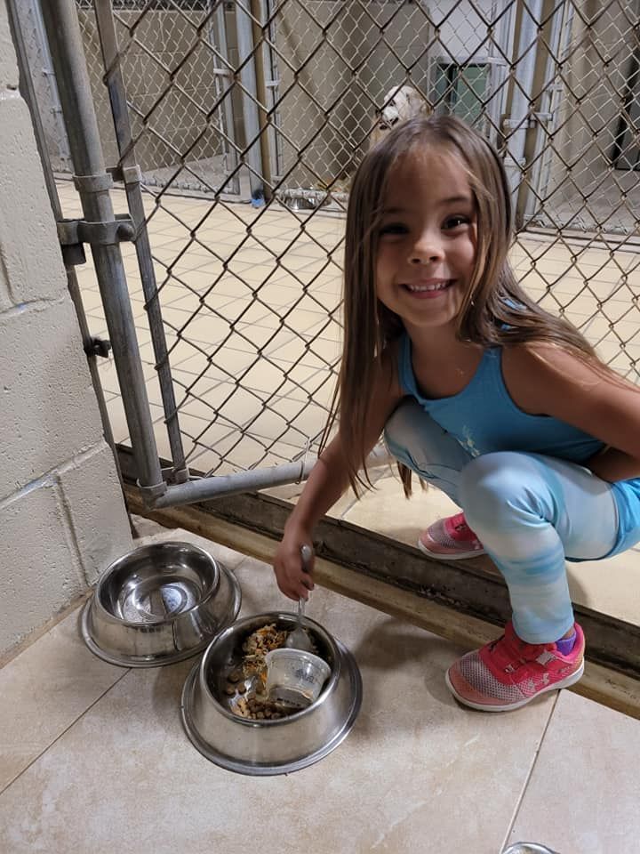 A young girl is smiles while crouching by a kennel serving dog food in a metal bowl