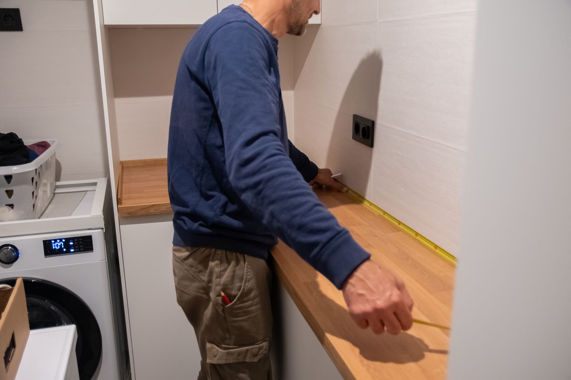 A person in a blue sweatshirt measuring a wooden countertop with a tape measure in a laundry room.