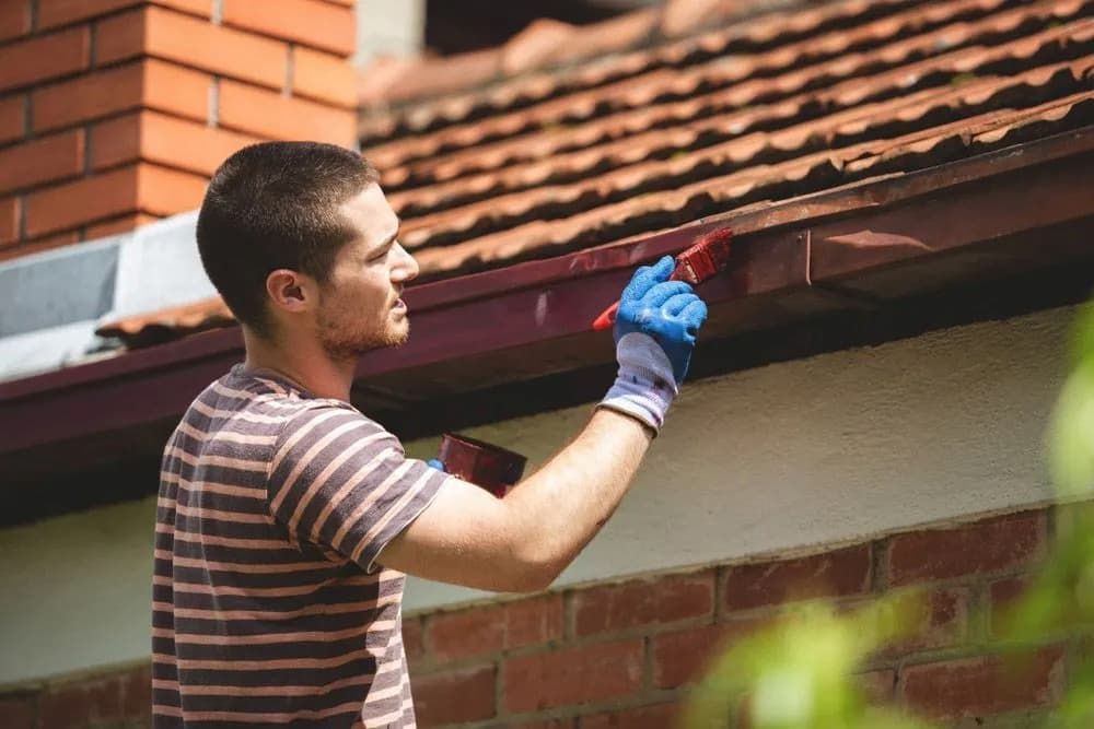 A Person Wearing a Striped Shirt and Blue Gloves Painting a House Eave With a Red Brush