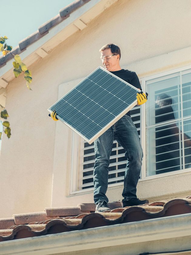 A man is standing on the roof of a house holding a solar panel.