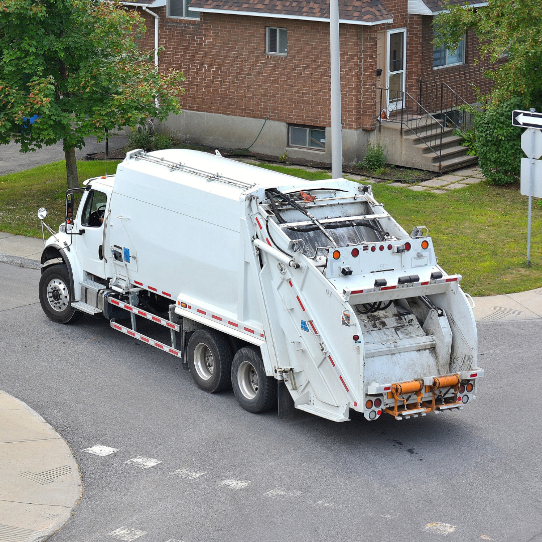 A white garbage truck is driving down a street in front of a brick house.