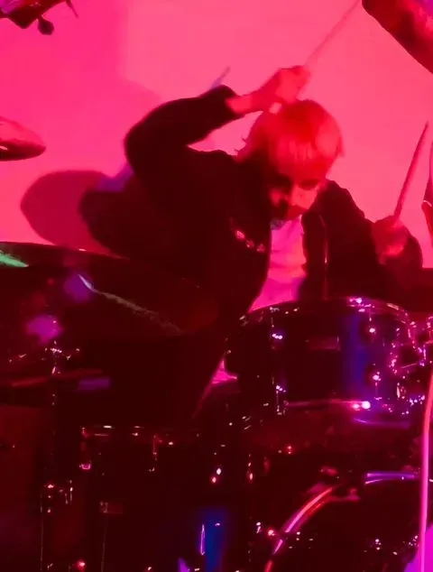 A person is playing drums on a stage in front of a pink background.