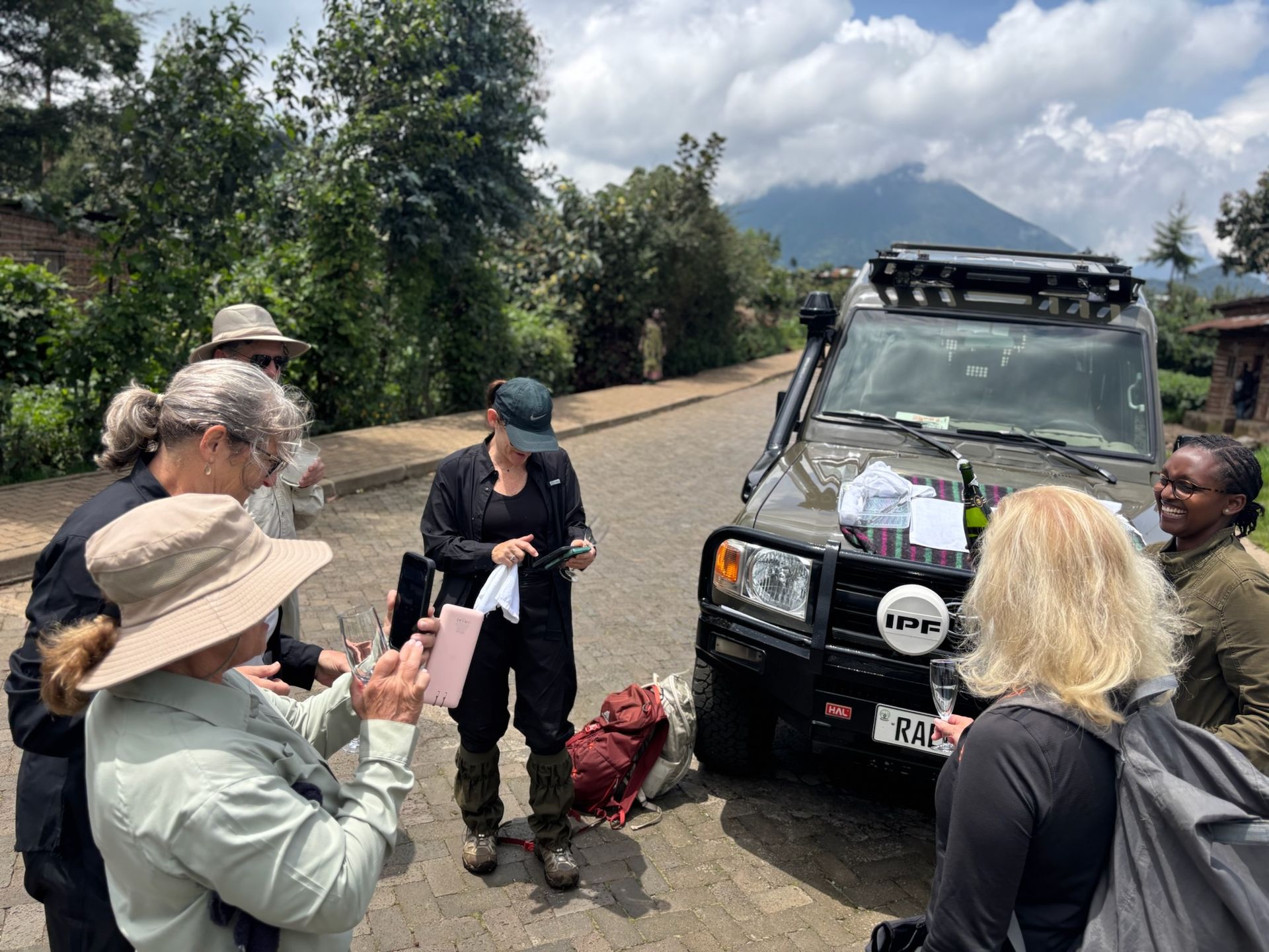 A group of people standing in a field in front of small, simple houses, with a terraced, green mountain in the background.