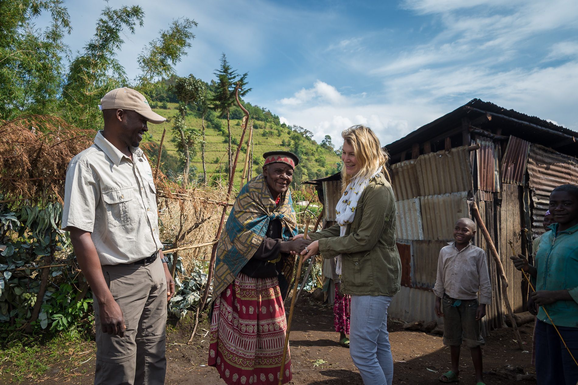 A person in a tan jacket shakes hands with a local resident wearing traditional patterned clothing in a rural setting.